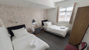 A hotel room with two beds, a desk, a chair, a mirror, and a window with curtains. - Mayfair Hotel (Skegness)