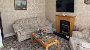 A living room with patterned wallpaper, a sofa, a coffee table with magazines, a fireplace, and a television. - Mayfair Hotel (Skegness)