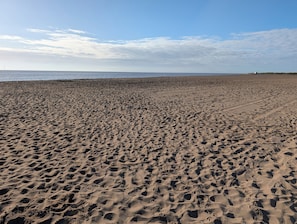 Ein sandiger Strand mit Fußabdrücken, einem klaren Himmel und einer entfernten Windkraftanlage.
