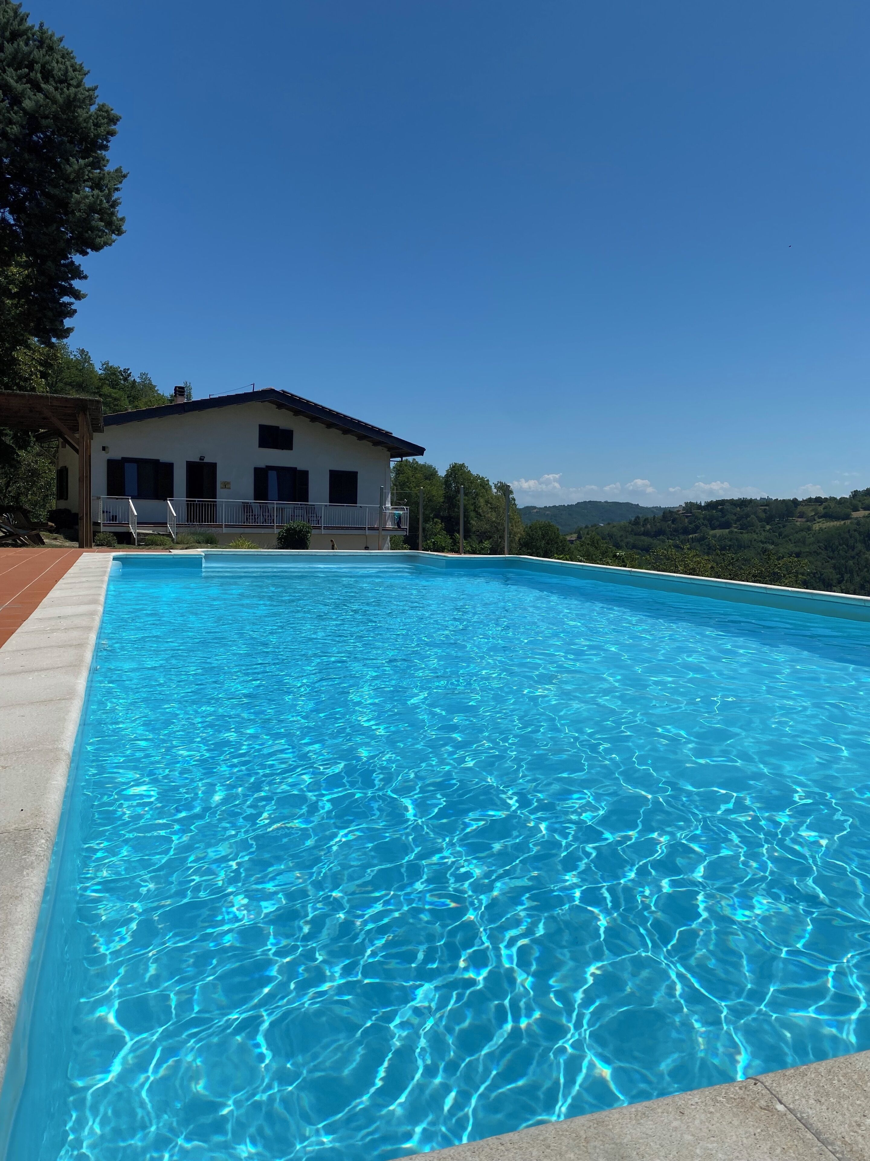 Une piscine rectangulaire à l’eau d’un bleu limpide, un bâtiment blanc aux volets sombres, et une vue sur des collines verdoyantes en arrière-plan.