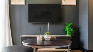 Une chambre moderne avec un téléviseur à écran plat fixé au mur, une table ronde en bois avec deux chaises, une petite plante en pot et un meuble en bois placé sous le téléviseur.
