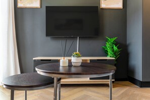 A modern room with a flat-screen TV mounted on the wall, a round wooden table with two chairs, a small potted plant, and a wooden cabinet below the TV. - Modern Studio | Nine Palms Century City (Cape Town)