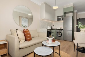 A compact living space with a sofa, a round mirror, a small table with a decorative item, a washing machine, and a refrigerator. - Charlie and the Chairman | Stylish Studio (Cape Town)
