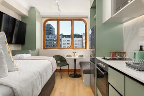Ein kleines Hotelzimmer mit Bett, Esstisch, Stuhl, Wandfernseher und Fenster mit Blick auf die Stadt.