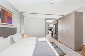 A modern hotel room with a bed, a desk, a chair, and a view into the kitchen area. - One Rosebank | Slow Luxury, Perfected (Johannesburg)