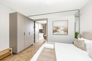 Ein modernes Schlafzimmer mit einem großen Bett, einem Nachttisch, einem Wandbild und Blick in den Küchenbereich.