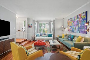 Ein Wohnzimmer mit Fernseher, gelben Sesseln, einer Couch, einem Couchtisch und einem farbenfrohen, abstrakten Gemälde.