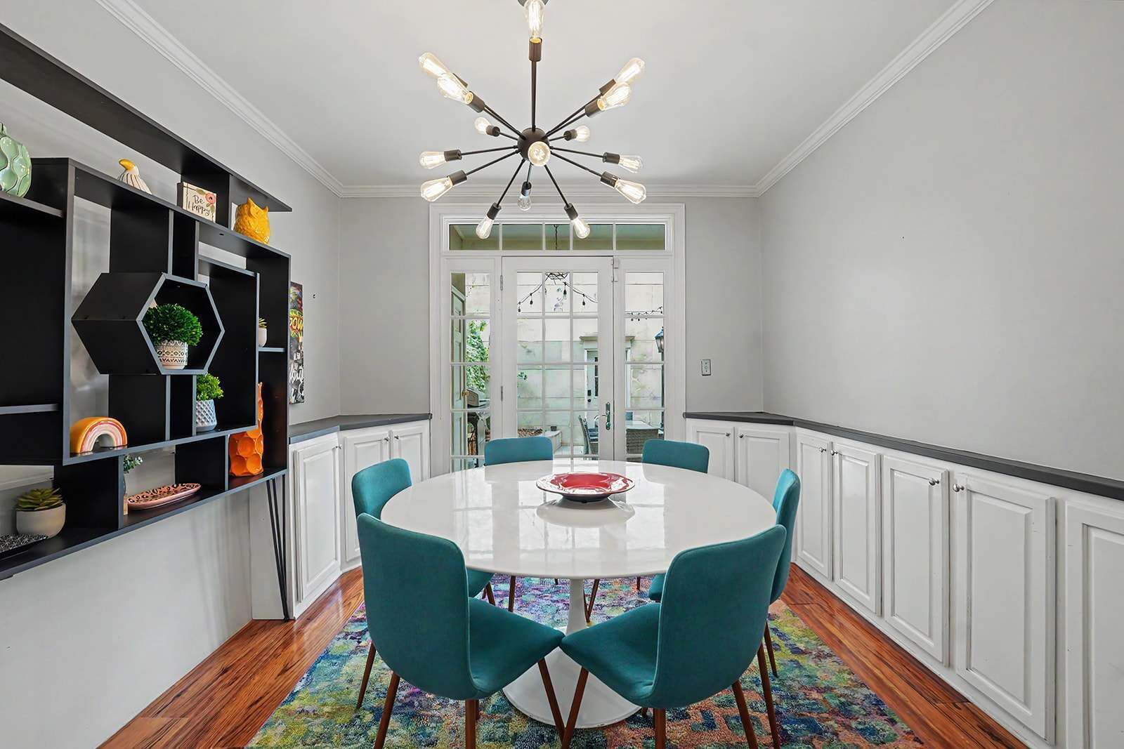 A dining area with a round table, teal chairs, a chandelier, and a colorful rug.