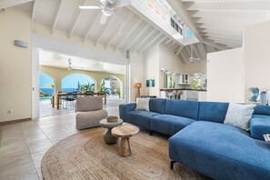 Un vaste espace de vie comprenant un canapé bleu, une table basse ronde et un coin repas offrant une vue sur l’océan.