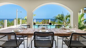 Uma sala de jantar com vista para o oceano, uma piscina e plantas tropicais.