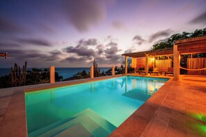 En swimmingpool med havudsigt, et overdækket loungeområde og en pergola med lyskæder.