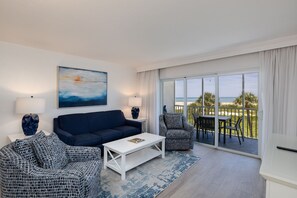Uma sala de estar moderna com sofá, mesa de centro e poltrona, um quadro na parede e um varanda com vista para o mar.