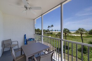 Varanda com mesa e cadeiras, ventilador de teto e vista para a praia e palmeiras.