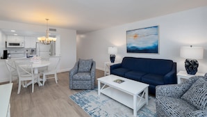 Ein modernes Wohnzimmer mit einem blauen Sofa, einem weißen Couchtisch, gemusterten Sesseln, einem Esstisch mit Stühlen, einer Küchenzeile mit weißen Schränken und einer Mikrowelle sowie einem Bild an der Wand.
