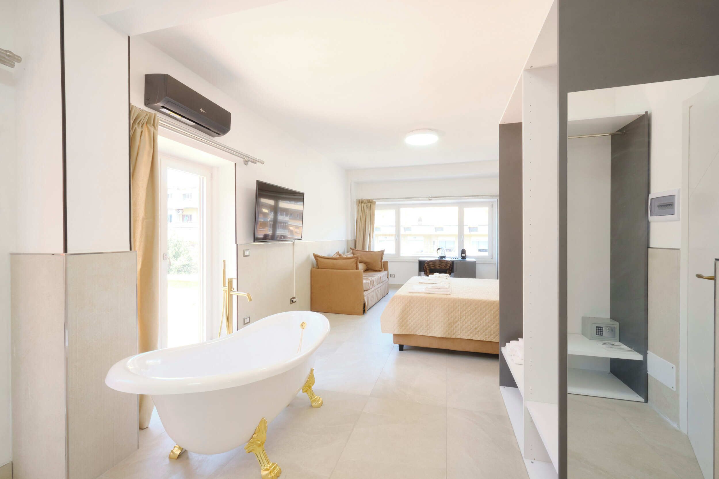 A modern hotel room with a bathtub, a bed, a desk, and a view of the outside.