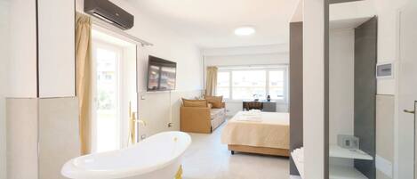 A modern hotel room with a bathtub, a bed, a desk, and a view of the outside.