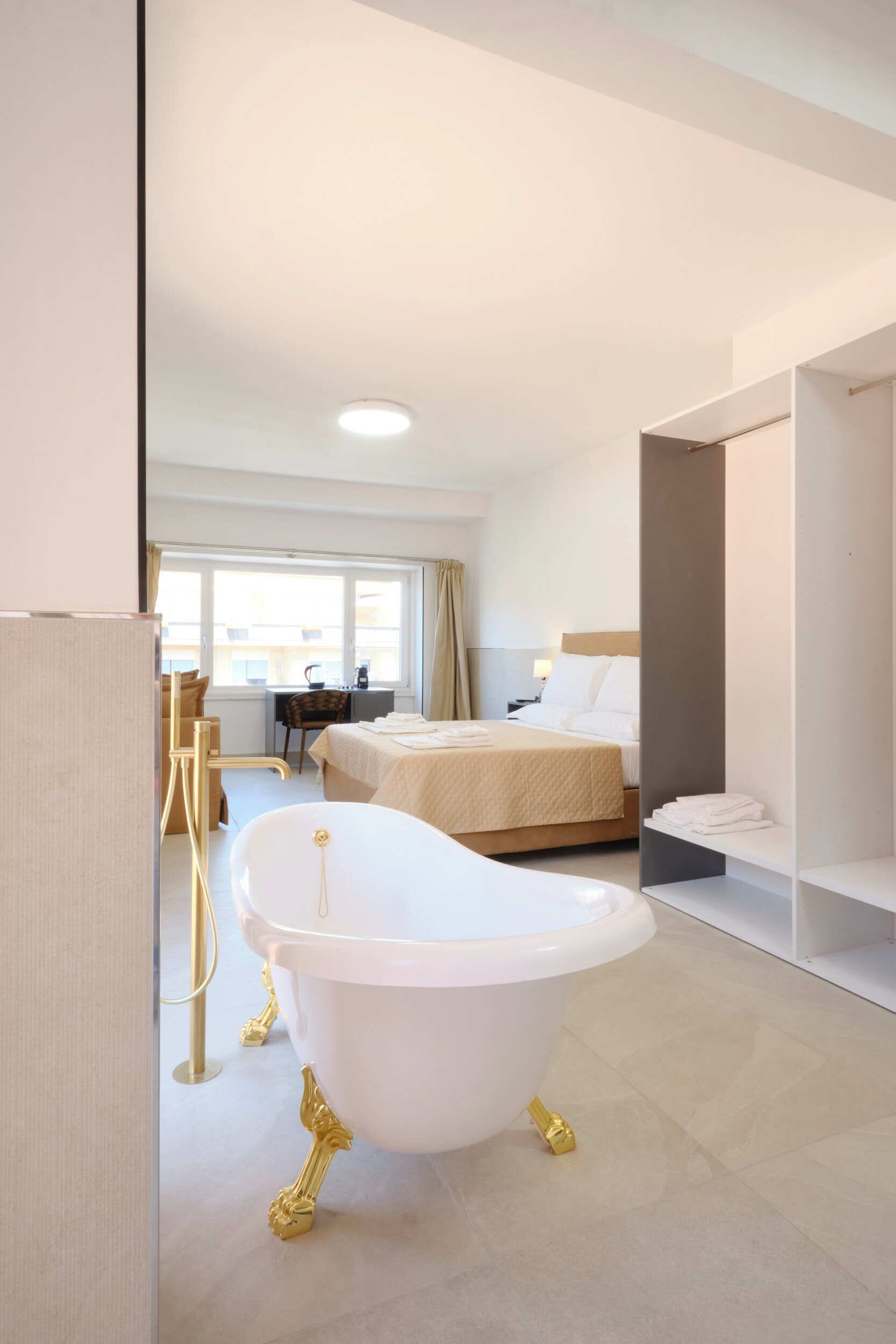 A modern hotel room with a bathtub, a bed, a desk, and a window.