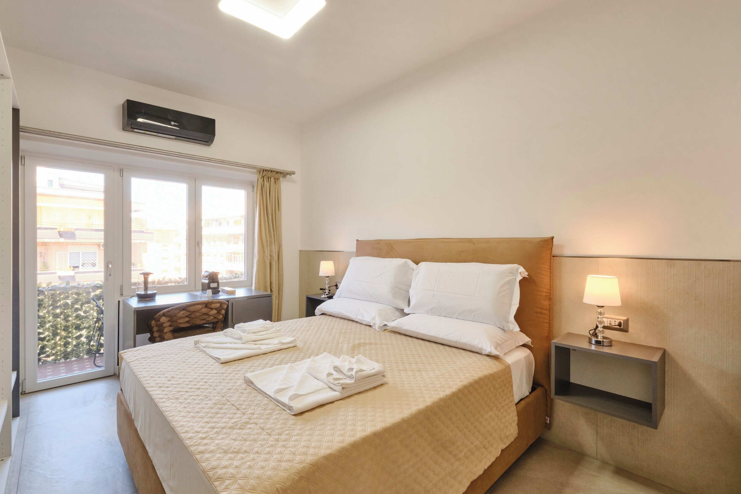 A hotel room with a bed, a desk, a chair, a nightstand with a lamp, and a window with curtains.
