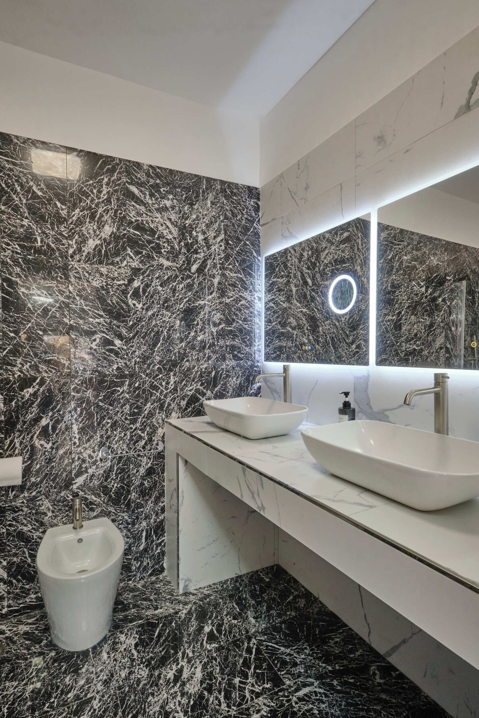 A modern bathroom with two sinks, a urinal, and a large mirror with a circular light.