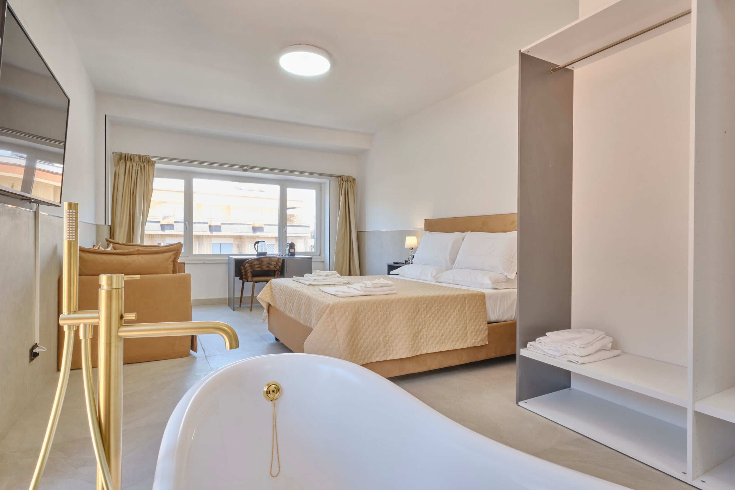 A modern hotel room with a large bed, a sitting area with a chair and table, a bathtub, and a wardrobe.