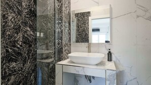 Un baño con lavamanos blanco, un espejo y una zona de ducha con paredes de mármol negro.
