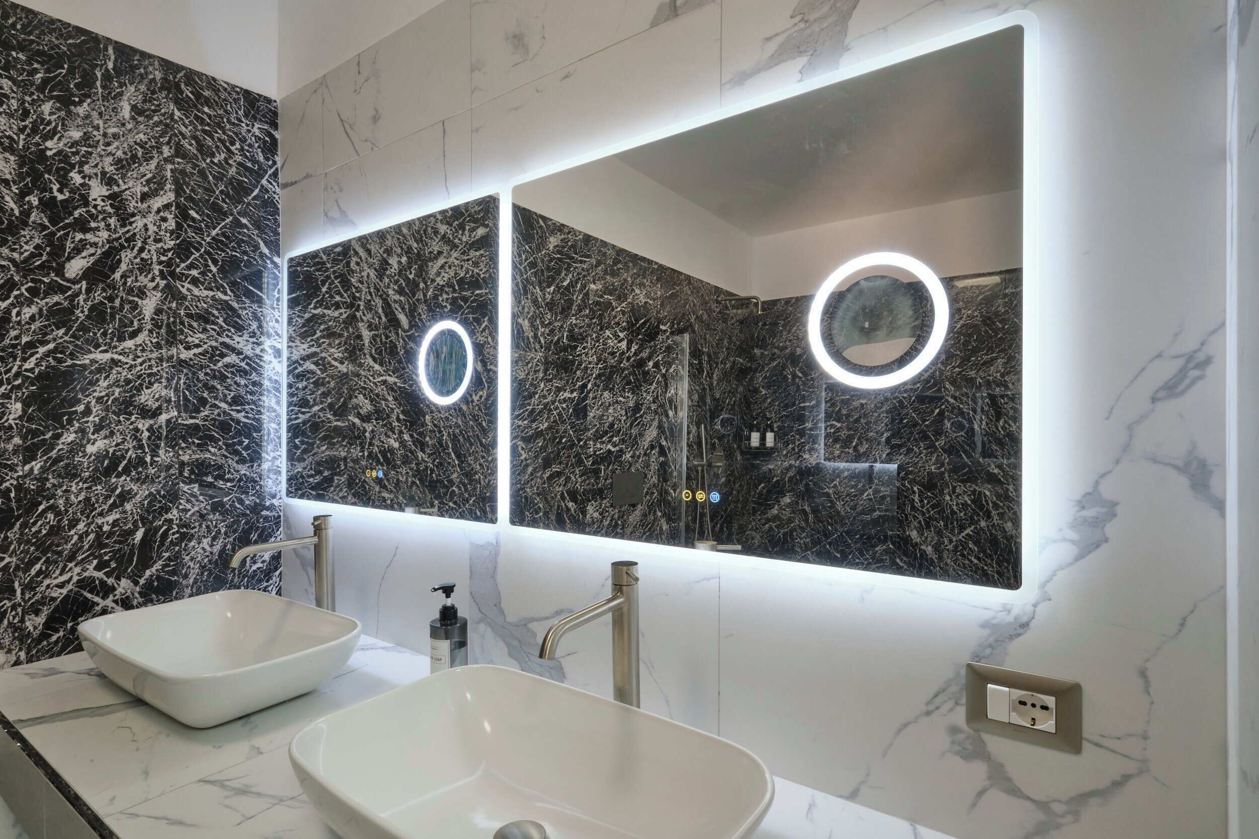 Une salle de bain avec deux lavabos, un grand miroir doté d’un éclairage intégré et des murs en marbre.