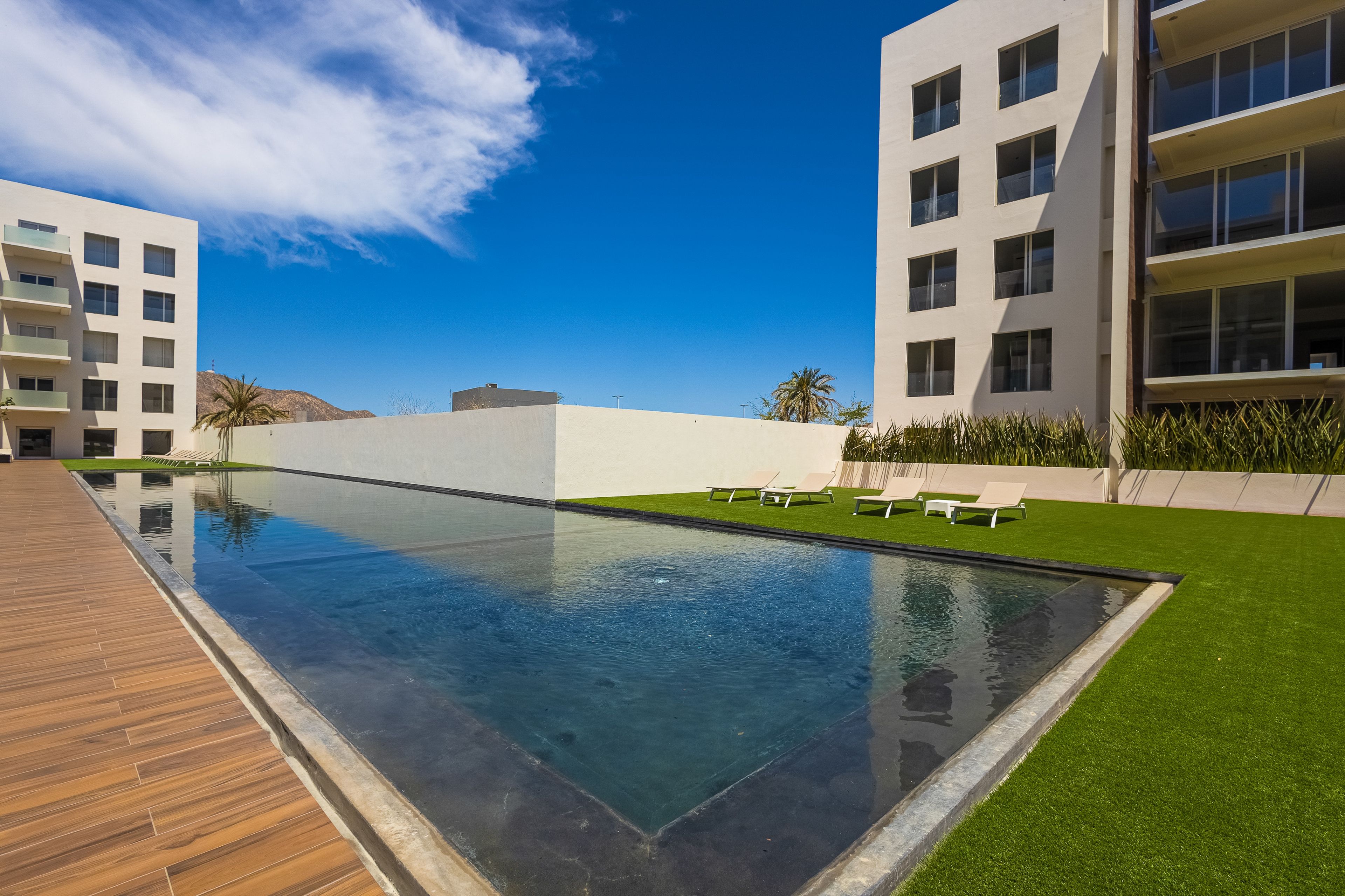 A modern pool area with a wooden deck, surrounded by white buildings and green grass.