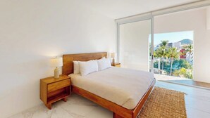 Un dormitorio con una cama grande, una mesita de noche, una lámpara y vistas a palmeras y edificios.