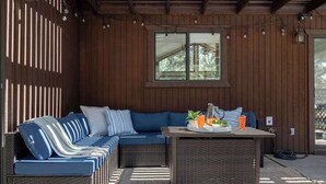 Ein überdachter Außenbereich mit Sitzgelegenheiten, einer Rattan-Couch, einem Tisch mit Getränken und einem Fenster mit Blick auf Bäume.
