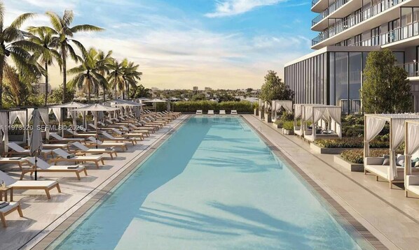 Una piscina con lettini e ombrelloni, circondata da palme e un edificio moderno.