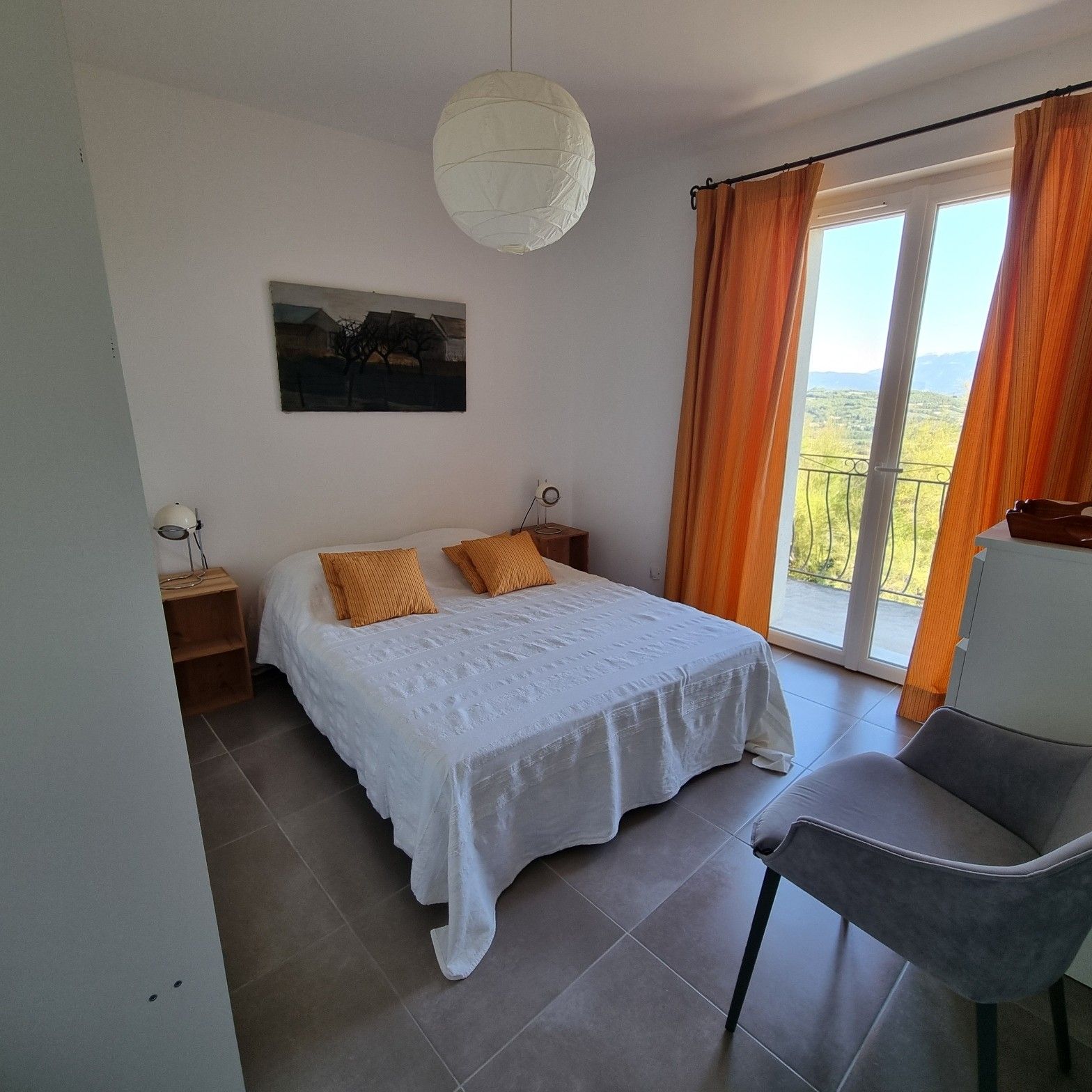 A bedroom with a bed, a chair, a nightstand, a lamp, a painting, and a view of the outdoors.