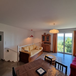 A living room with a sofa, a dining table, a cabinet, and a view through a large window. - Apartment 'La Roseraie Des Côtes' with Private Pool, Private Garden and Wi-Fi (Vinsobres)