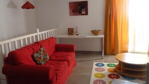 A red sofa with a patterned pillow, a white table with a bowl and a lamp, and a colorful mat with buttons. - Apartment 'La Roseraie Des Côtes' with Private Pool, Private Garden and Wi-Fi (Vinsobres)