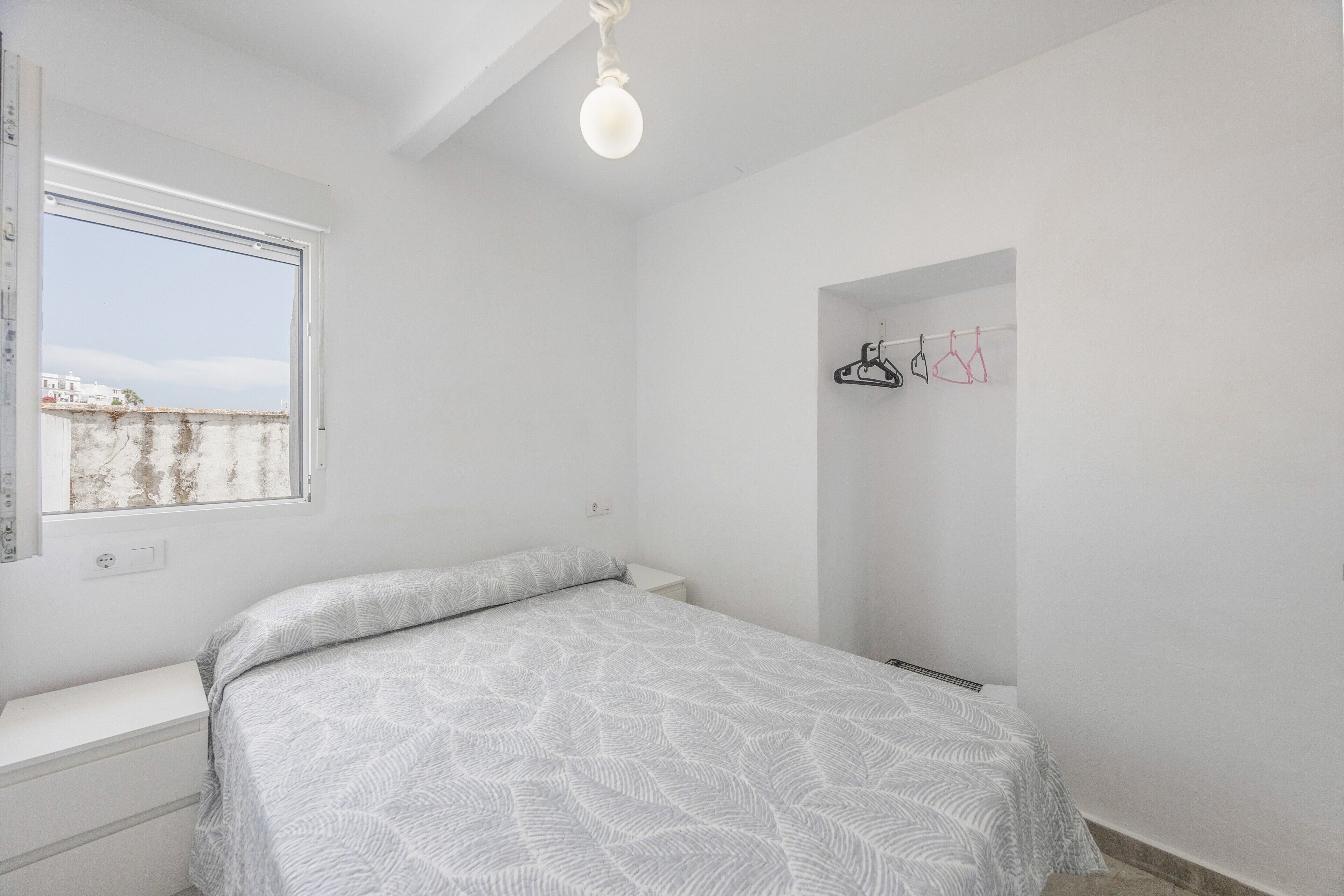 A bedroom with a bed, a window, a hanging light fixture, and a wall-mounted coat rack.