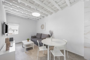 A modern living room with a white sofa, a round table with chairs, a flat-screen TV mounted on the wall, and a window with a view. - 'La Joyita Del Centro' Apartment with Shared Garden and Wi-Fi (Vejer de la Frontera)