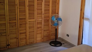 Une chambre avec une armoire en bois, un ventilateur sur pied et un lit.