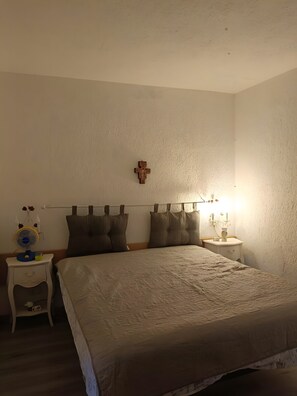 Une chambre à coucher comprenant un lit, une table de chevet, une commode, un crucifix fixé au mur et un ventilateur fixé au mur.