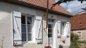 Une maison aux murs blancs et au toit recouvert de tuiles rouges, un jardin avec une chaise bleue et un pot de fleurs contenant des fleurs rouges.