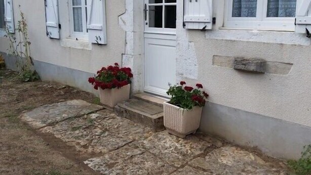 Un bâtiment à l’extérieur blanc, avec une porte et des fenêtres dotées de volets. Devant la porte, on trouve des plantes en pot ornées de fleurs rouges.