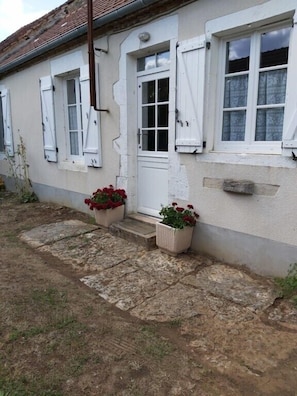 A building with a white exterior, a door, and windows with shutters. There are potted plants with red flowers in front of the door.