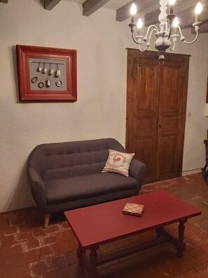 Een kamer met een donkere bank, een rode tafel, een houten deur en een kroonluchter.