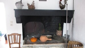 Une cheminée en pierre avec un manteau noir, entourée d’une décoration rustique comprenant un fauteuil en osier, une chaise en bois et des objets décoratifs.