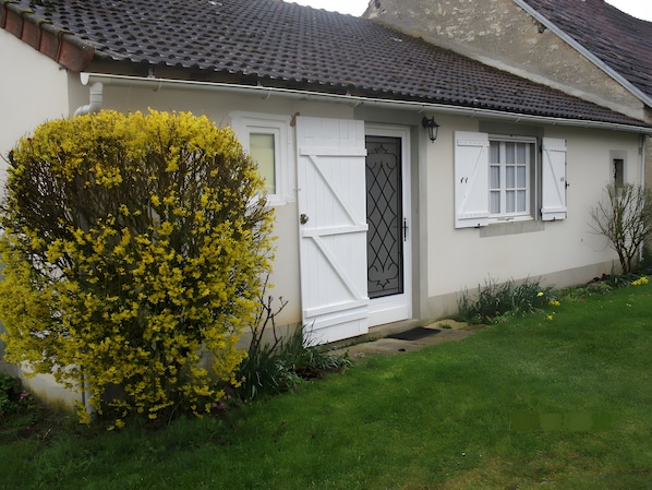 Une maison à l’extérieur blanc, avec un toit en tuiles sombres, une porte blanche et un portail en métal noir, ainsi qu’un petit jardin agrémenté d’arbustes à fleurs jaunes.
