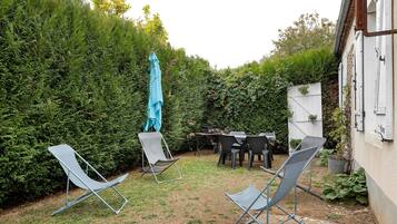 Een achtertuin met een tafel, stoelen, een blauwe parasol en een haag.