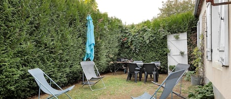 Um quintal com mesa, cadeiras, um guarda-sol azul e um arbusto.