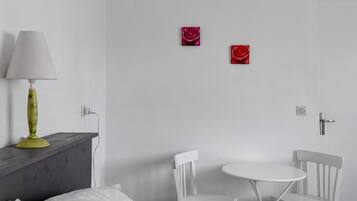 Una habitación con una cama, una mesita de noche con lámpara, dos sillas blancas, una pequeña mesa redonda y dos cuadros de rosas rojas en la pared.