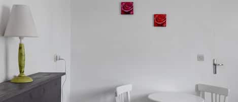 Una habitación con una cama, una mesita de noche con lámpara, dos sillas blancas, una pequeña mesa redonda y dos cuadros de rosas rojas en la pared.