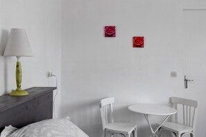Una habitación con una cama, una mesita de noche con lámpara, dos sillas blancas, una pequeña mesa redonda y dos cuadros de rosas rojas en la pared.