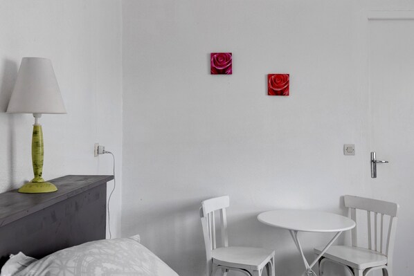 A room with a bed, a nightstand with a lamp, two white chairs, a small round table, and two red rose paintings on the wall.