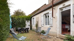A rustic outdoor seating area with metal chairs and a table, surrounded by a garden and a building with a tiled roof.